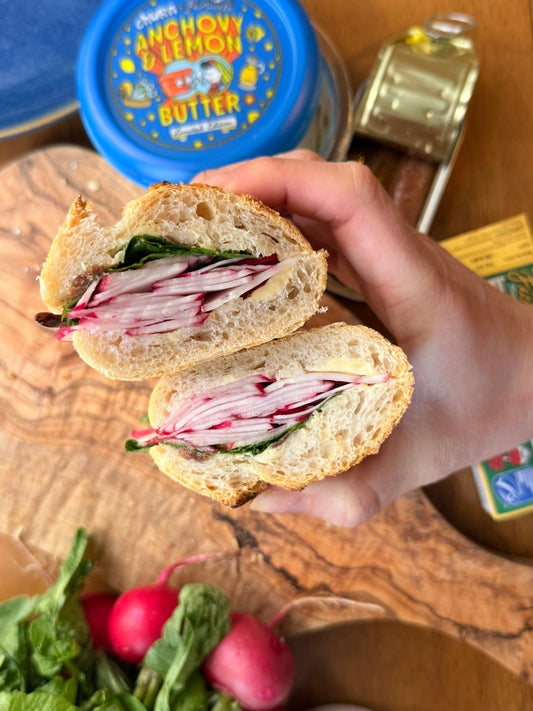 Hand holding radish and greens sandwich with anchovy lemon butter on wooden board, open butter container behind.
