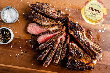 Sliced medium-rare steak with garlic & shallot butter on a wooden board, served with salt and pepper.