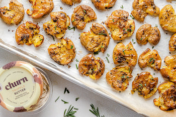 Crispy smashed roasted potatoes on a baking sheet with herbs, paired with Churn Truffle Butter.