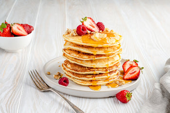 Stack of pancakes drizzled with syrup, topped with butter, berries, and walnuts, served with fresh strawberries and fork.