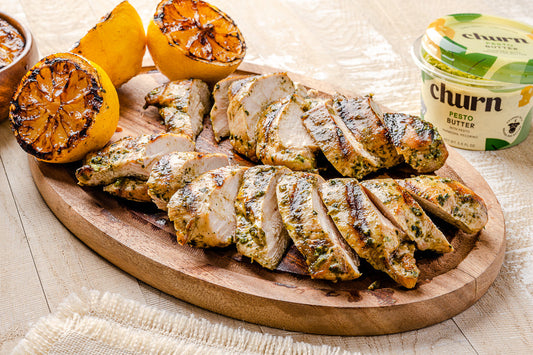 Sliced grilled pesto chicken on a wooden board with charred lemon halves and a tub of Churn Pesto Butter.