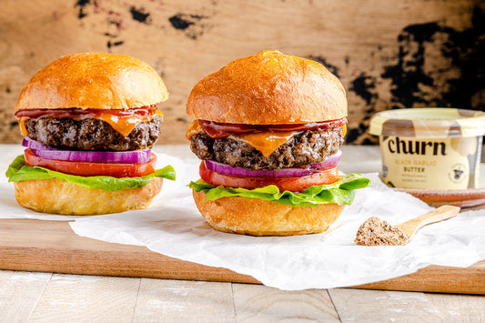 Two beef burgers with cheddar, onion, tomato, and lettuce on brioche buns beside Churn Black Garlic Butter.