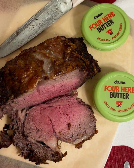 Sliced medium-rare roast beef with a seared crust on a cutting board alongside two containers of Churn Four Herb Butter.