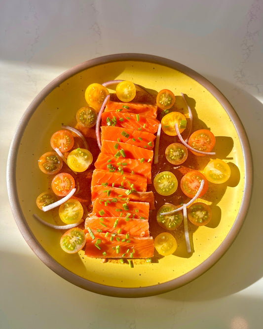Sliced cured salmon with chives, halved cherry tomatoes, and red onion on a yellow speckled plate in sunlight.