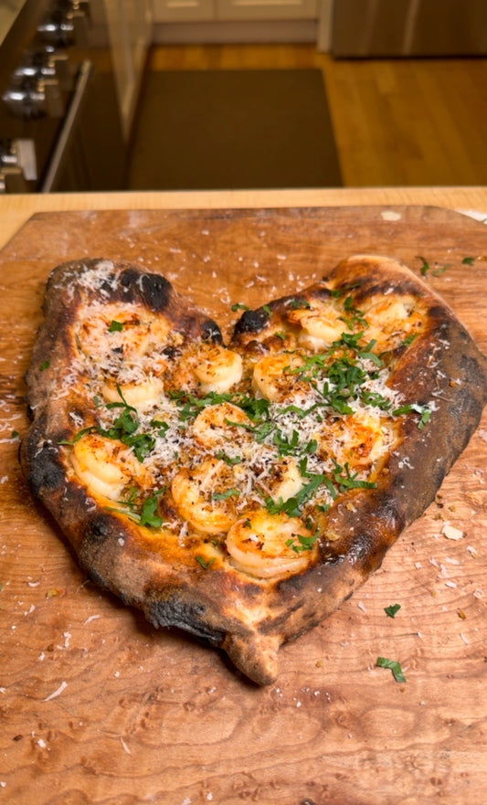 Charred rustic pizza topped with shrimp, grated cheese, and fresh herbs on a wooden board.