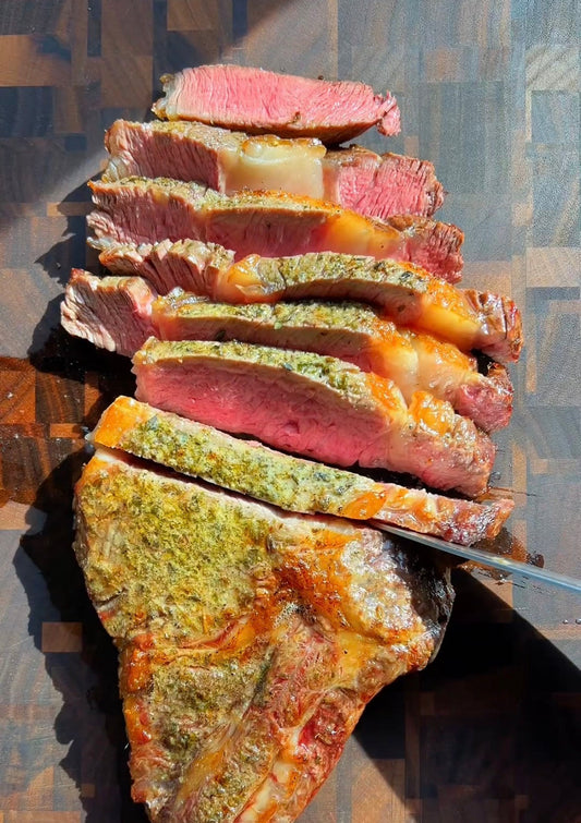 Sliced medium-rare ribeye steak with herb butter crust on a wooden cutting board in natural sunlight.