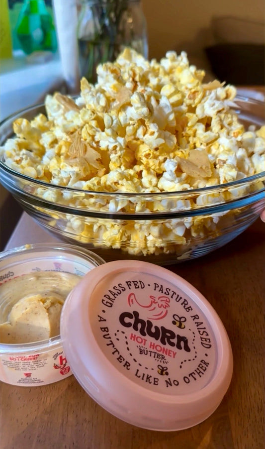 A large glass bowl of buttered popcorn with almonds alongside an open container of Churn Hot Honey Butter.