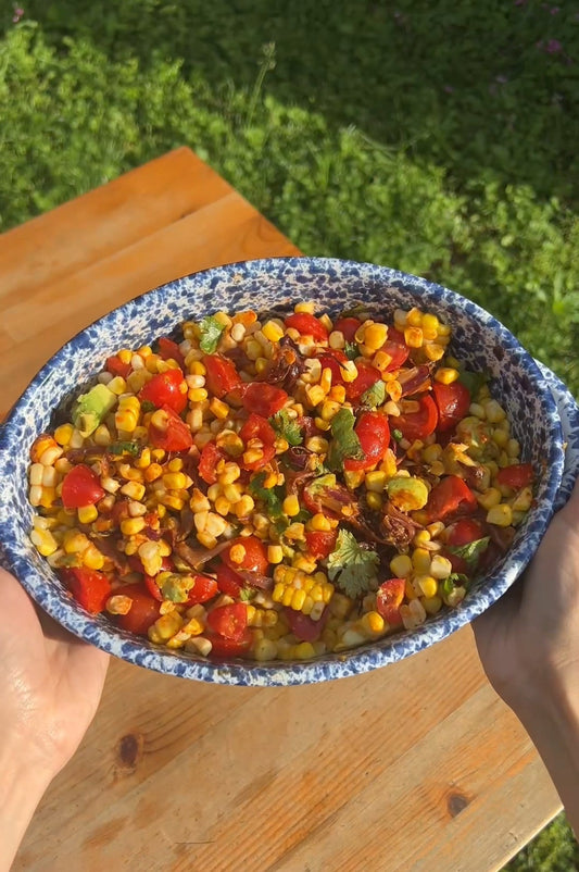 Hands holding a blue speckled bowl of corn salad with cherry tomatoes, avocado, and cilantro outdoors.
