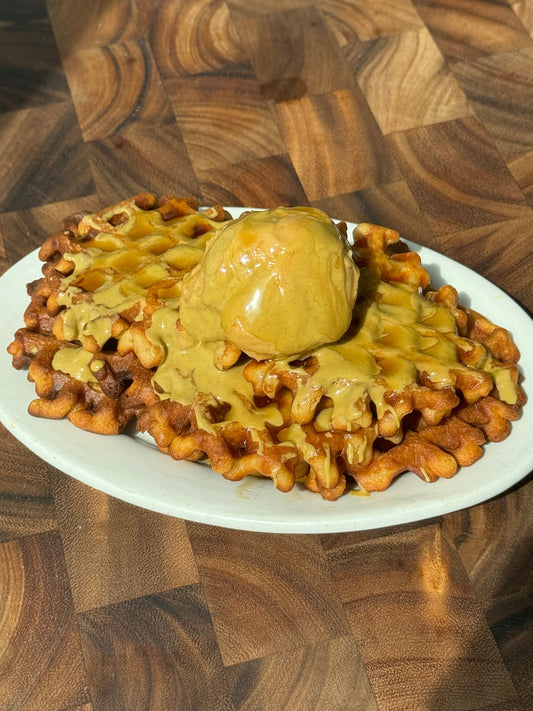A stack of waffles topped with ice cream and golden sauce on an oval white plate on a wooden surface.
