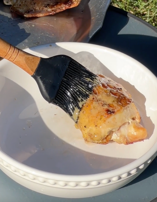 A silicone basting brush applying melted butter to a grilled chicken thigh in a white bowl outdoors.