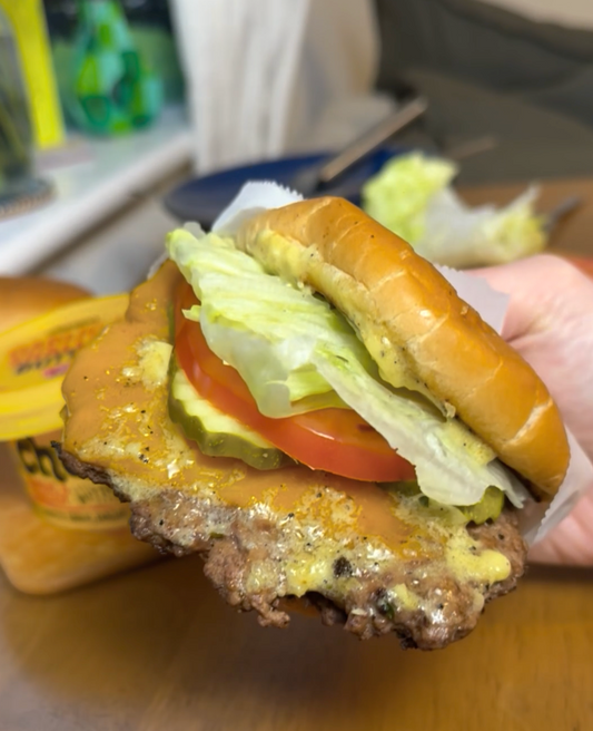 A hand holding a smash burger with melted cheese, pickles, tomato, and lettuce on a brioche bun.