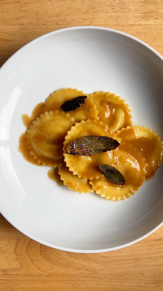 Plate of ravioli in brown butter sauce topped with crispy sage leaves on a wooden table.
