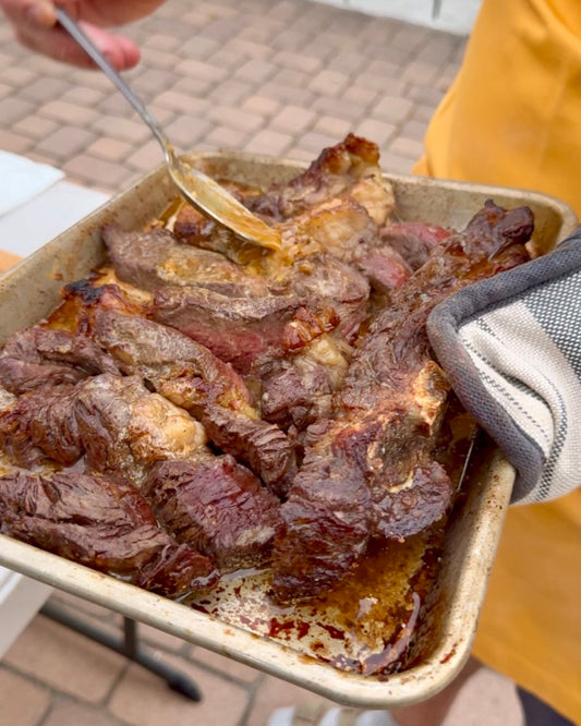 Person outdoors holding tray of cooked steaks, spooning sauce over meat, wearing an oven mitt.