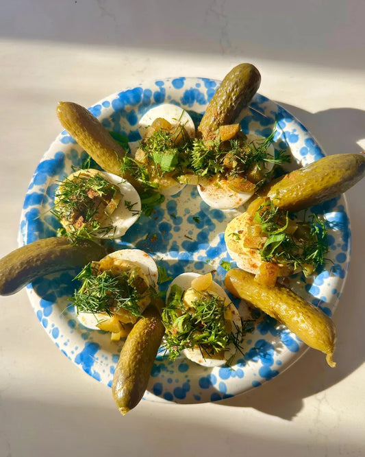 Deviled eggs topped with fresh dill, paprika, and pickles served on a blue speckled plate in natural sunlight.